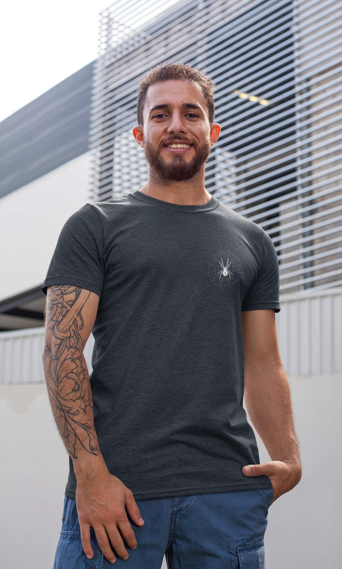 Man with tattoos standing with hand in pocket wearing tee of a black widow on a web over the left chest area